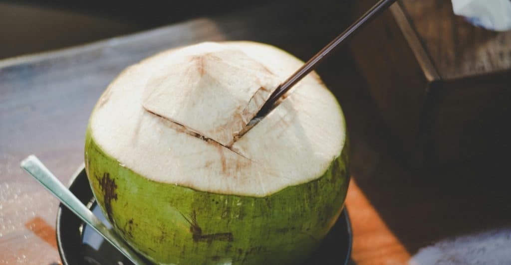 A whole green coconut with its top cut open and a straw inserted, sits on a black saucer with a spoon.