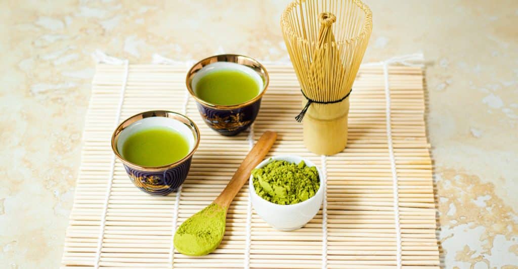 Two small cups of green tea, a whisk, and a bowl of matcha powder on a bamboo mat.