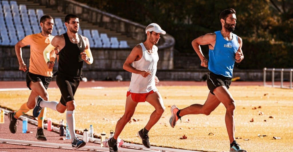 Four men wearing athletic wear are running together on a track in an outdoor stadium.