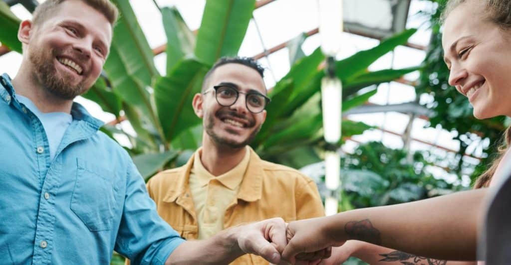 Three people are shown connecting in a friendly fist bump in a bright, plant-filled setting.