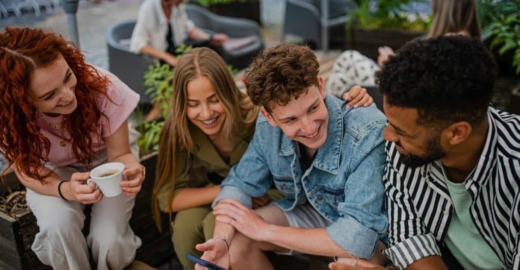 A diverse group of five friends sitting closely together outdoors, looking at a smartphone and smiling.