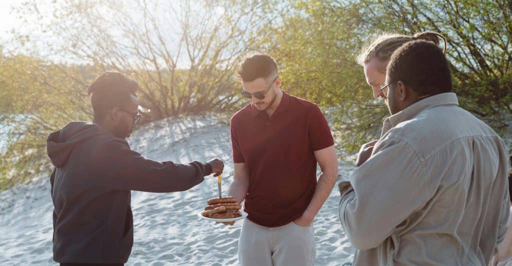 A group of people gathered around a barbecue grill on a sandy outdoor setting.