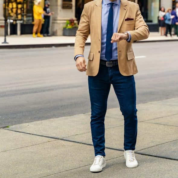 A man wearing a tan blazer, blue shirt, dark jeans, and white sneakers checks his wristwatch on a city sidewalk.