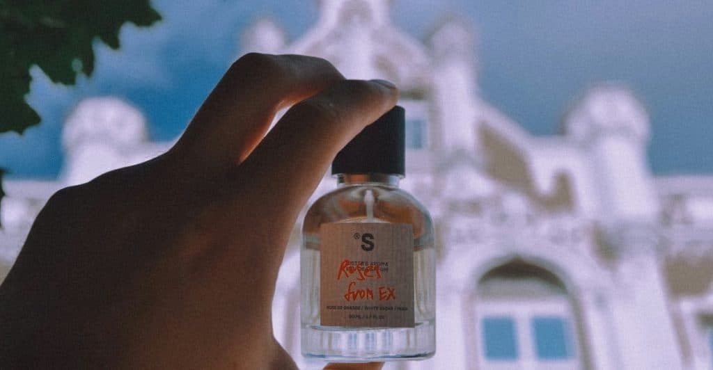 A hand holding a small fragrance bottle with a building in the blurred background.