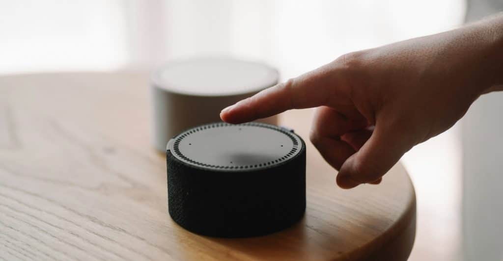 A hand reaching to touch the top of a voice assistant speaker on a wooden surface, illustrating interaction with smart home devices.