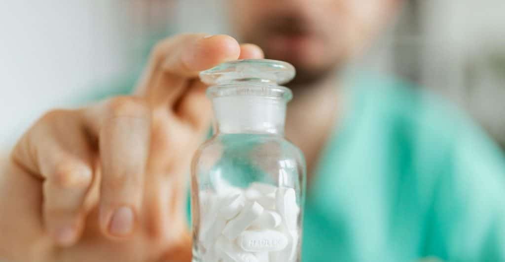  A person in a green shirt holds out a clear jar filled with white pills.