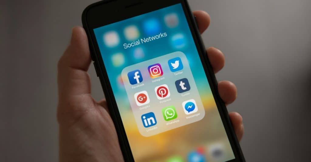 A hand holding a smartphone displaying icons for various social networking applications.