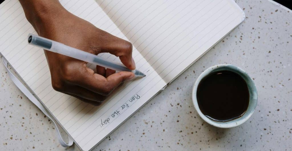 A person's hand writing "Plan for the day" in a notebook next to a cup of coffee.