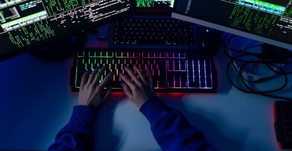 A person's hands typing on a brightly lit keyboard in front of computer screens.