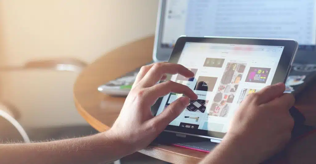 Hands are using a tablet to browse content on a table with a laptop in the background.