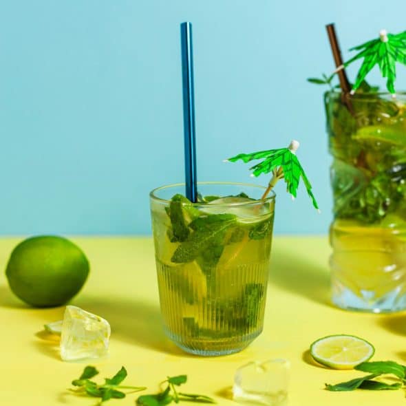 Two herbal drinks with lime and mint, ice cubes, and a blue straw on a yellow and blue background.