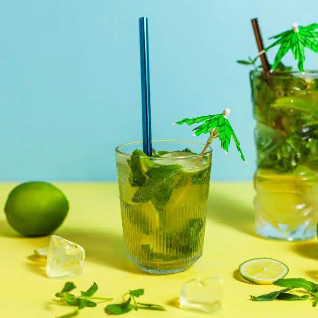 Two herbal drinks with lime and mint, ice cubes, and a blue straw on a yellow and blue background.