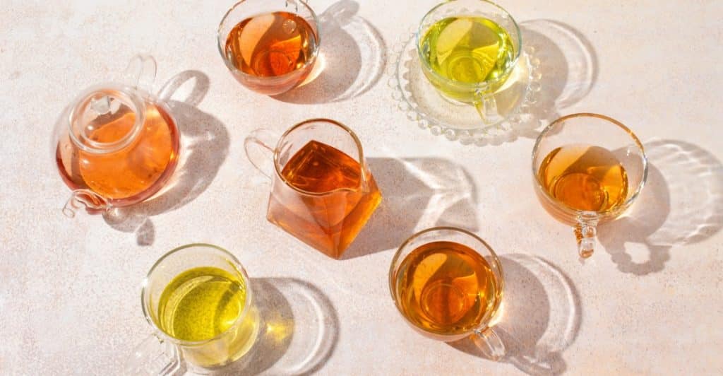 Several glass teacups and teapots filled with various shades of herbal tea arranged on a light-colored surface.