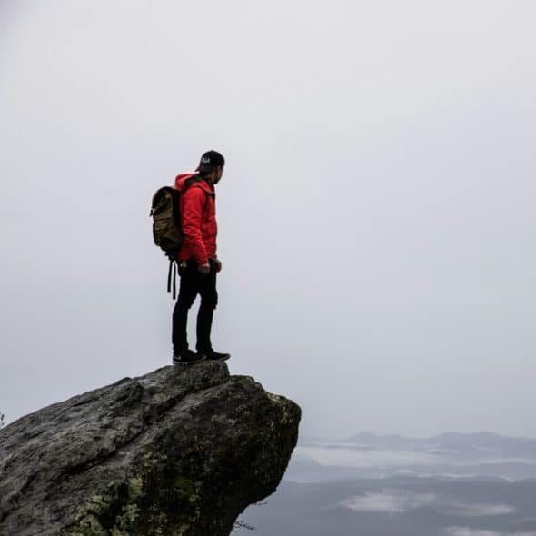 A person with a backpack standing on the edge of a rocky peak, looking out over a misty landscape.