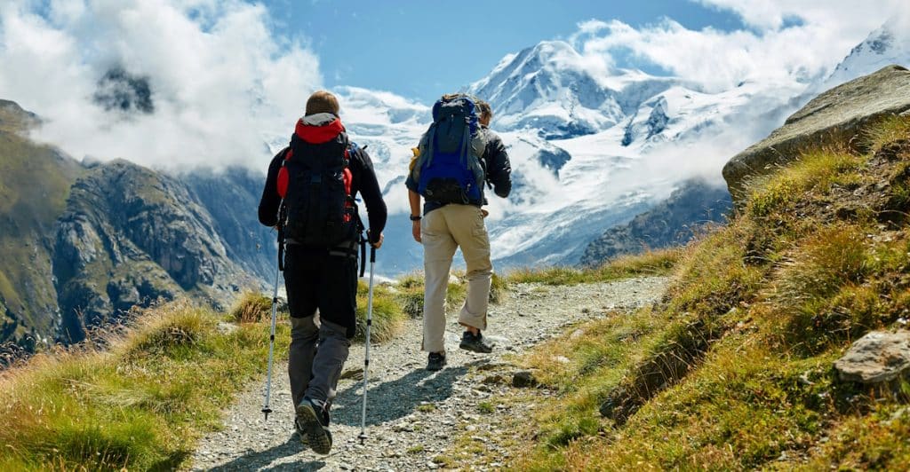 Two hikers with backpacks and hiking poles walking up a rocky trail in a mountainous landscape with a blue sky and clouds.