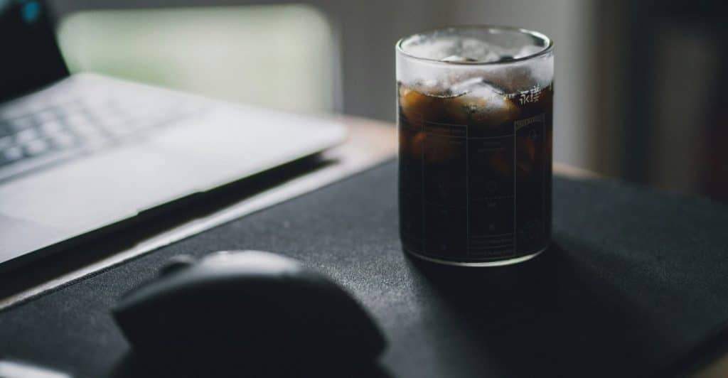 A glass of iced coffee sits on a desk next to a laptop and computer mouse.