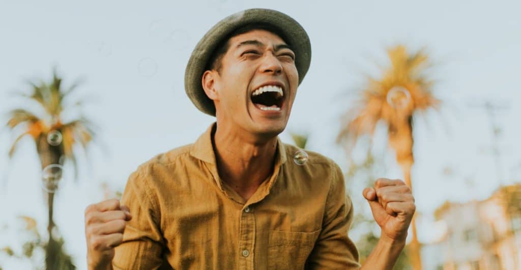 A person with a joyful expression, wearing a brimmed hat and a light brown shirt, with fists clenched, standing outdoors against a bright sky and blurred trees.