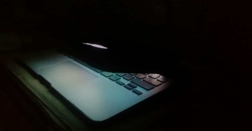 A laptop with partially closed lid, casting a blueish light on the keyboard and a small portion of the surrounding area.