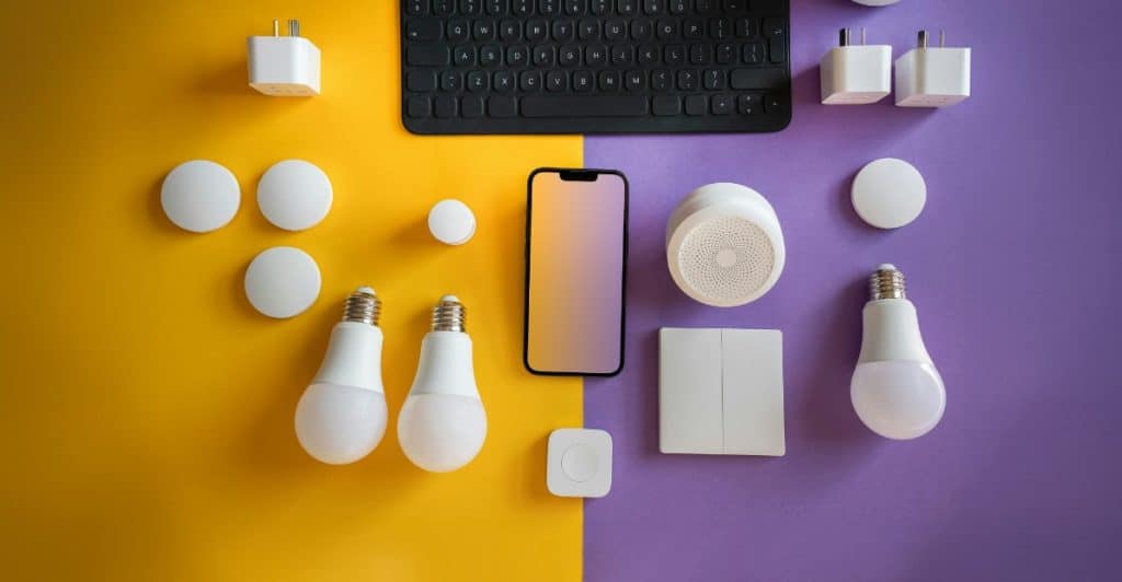 A flat lay arrangement of various smart home devices including a tablet, light bulbs, sensors, and a security camera on a colorful background, representing home automation.