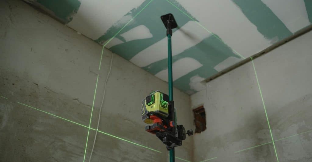 A laser level projects bright green horizontal and vertical lines onto the corner of a white room under construction.