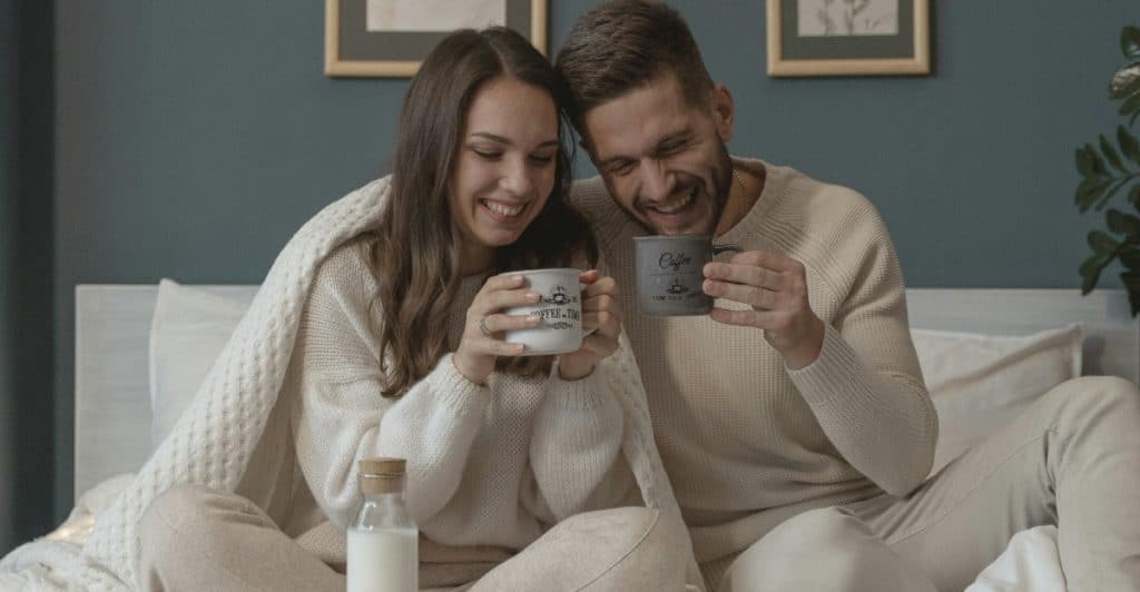 A couple wrapped in a blanket sitting in bed, laughing, and holding matching mugs, with a breakfast tray with a bottle of milk on the bed.