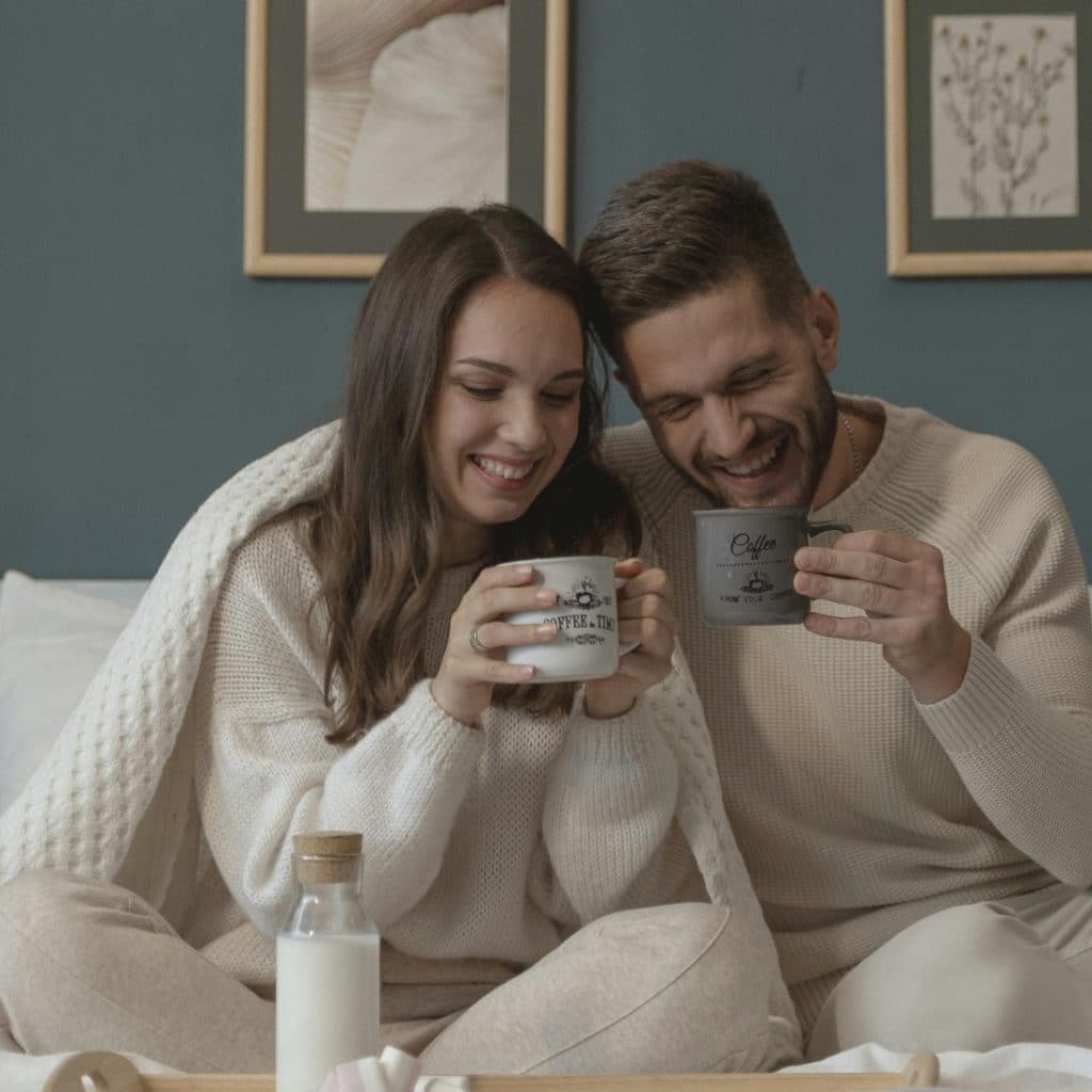A couple wrapped in a blanket  sitting in bed, laughing, and holding matching mugs, with a breakfast tray with a bottle of milk on the bed.