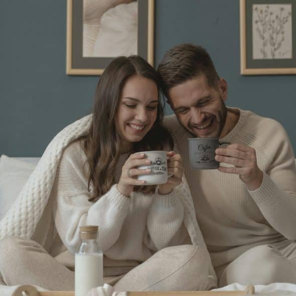 A couple wrapped in a blanket sitting in bed, laughing, and holding matching mugs, with a breakfast tray with a bottle of milk on the bed.