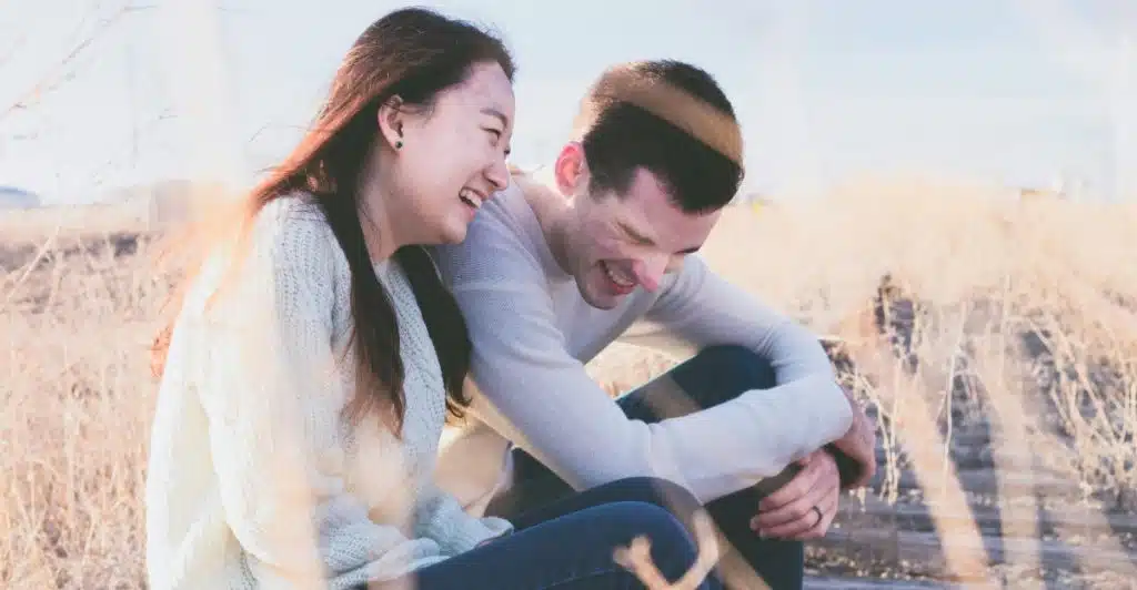 Couple sitting close together outdoors in a field of dry grass and laughing.