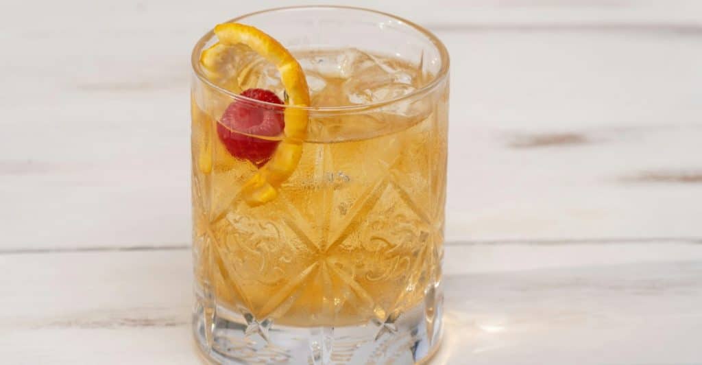 Lemonade with HoneyAlt-Text: A glass of lemonade and honey with ice, garnished with an orange peel twist and a raspberry, on a white wooden surface.