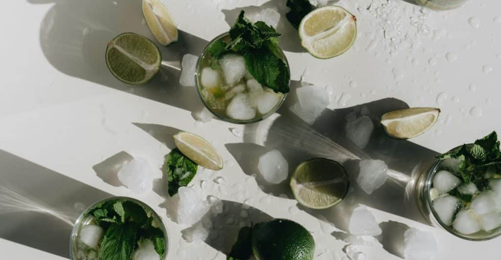 An overhead shot of several glasses with lime, mint, and ice, scattered on a wet white surface.