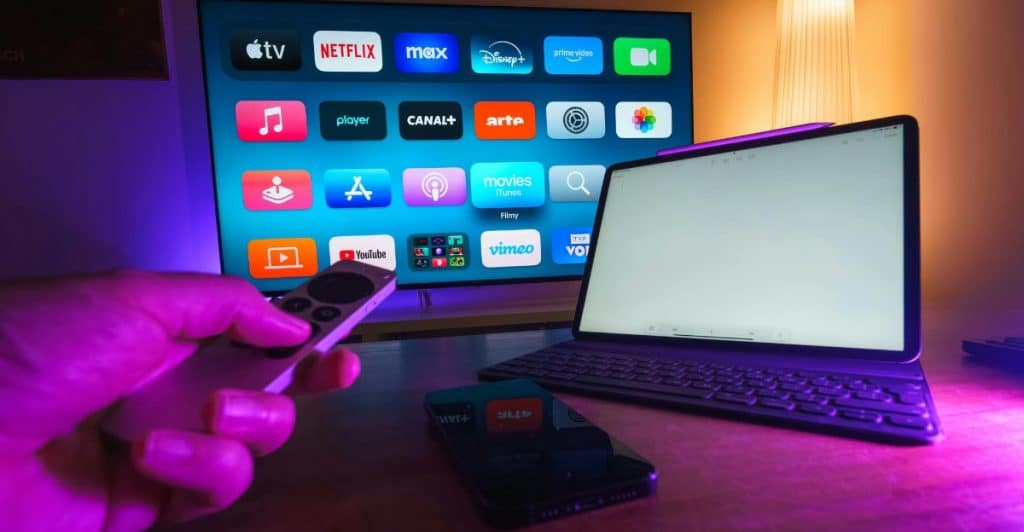 A hand holding a TV remote in front of a television screen displaying streaming service app icons, with a laptop and phone on the table.