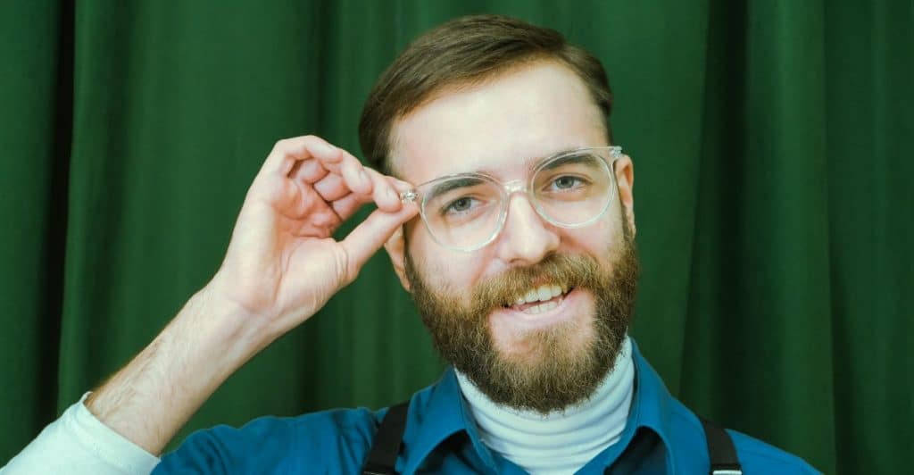 A smiling man with a beard and glasses adjusting his eyewear against a green background, suggesting the use of blue light filters or glasses.