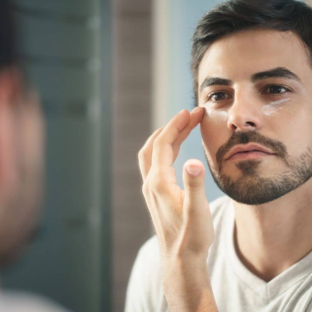 Bearded man applying eye cream while looking in a mirror