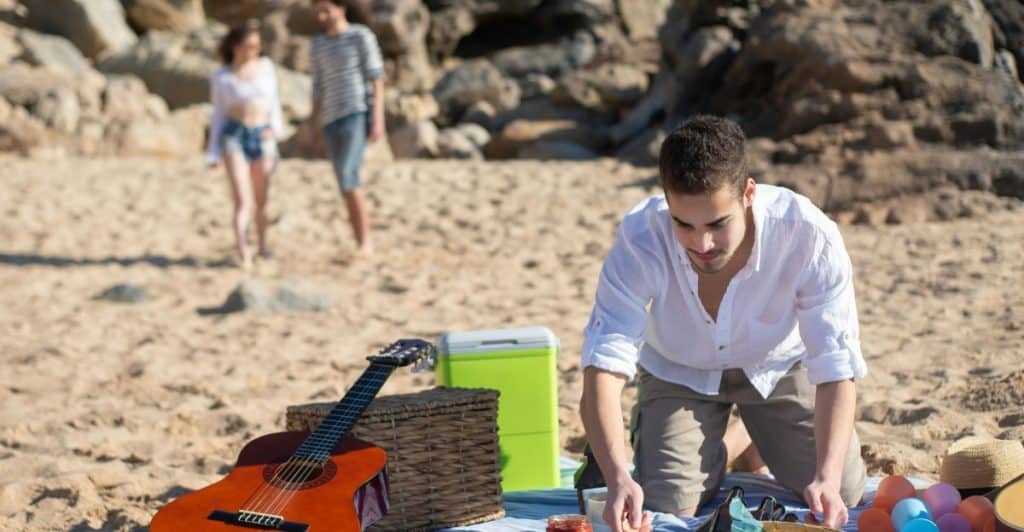 A man kneeling on a picnic blanket on a beach, arranging food, with a couple walking  in the background.
