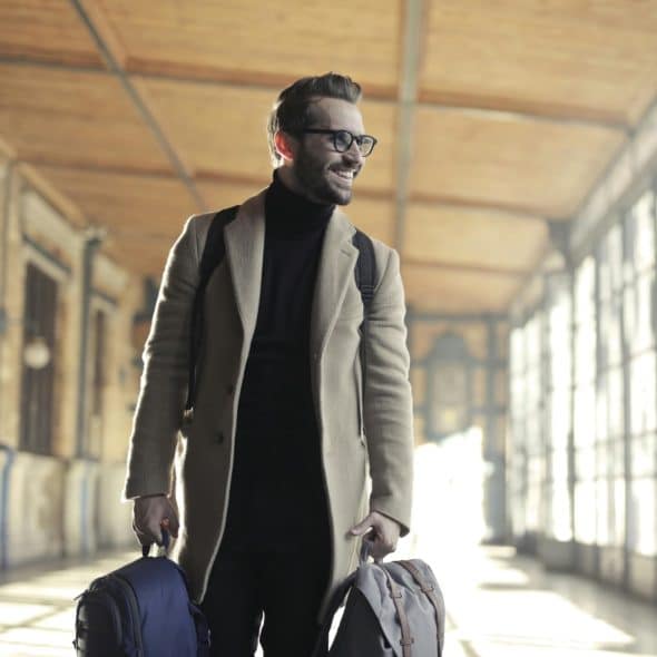 A smiling man in a tan overcoat, black turtleneck, and glasses walking through a bright, windowed hall, carrying a backpack on his back and two in his hands.