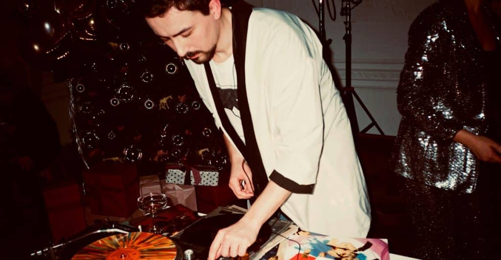 A man DJing with vinyl records at an event, with a reflective, sequined figure partially visible in the background.
