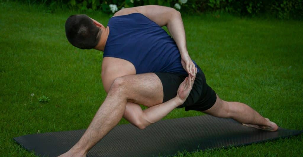  A man doing a seated yoga pose on a black mat in the grass, twisting his torso to the side, with one hand on the mat and the other reaching behind him.