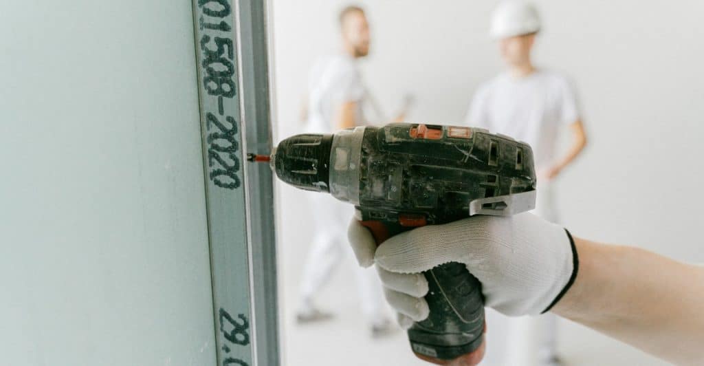 Close-up of a hand in a white glove holding a cordless drill against a drywall sheet marked with numbers.