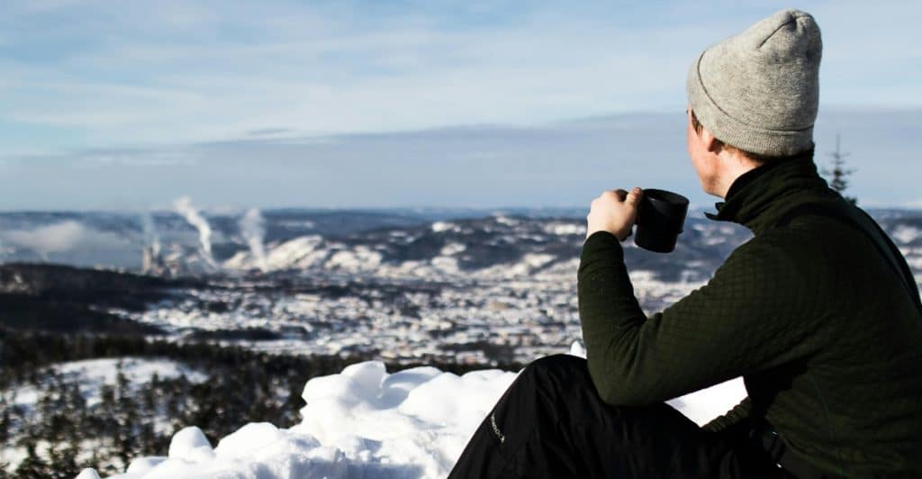 A person wearing a beanie and warm clothing sits on a snowy hill drinking from a dark mug, overlooking a town.
