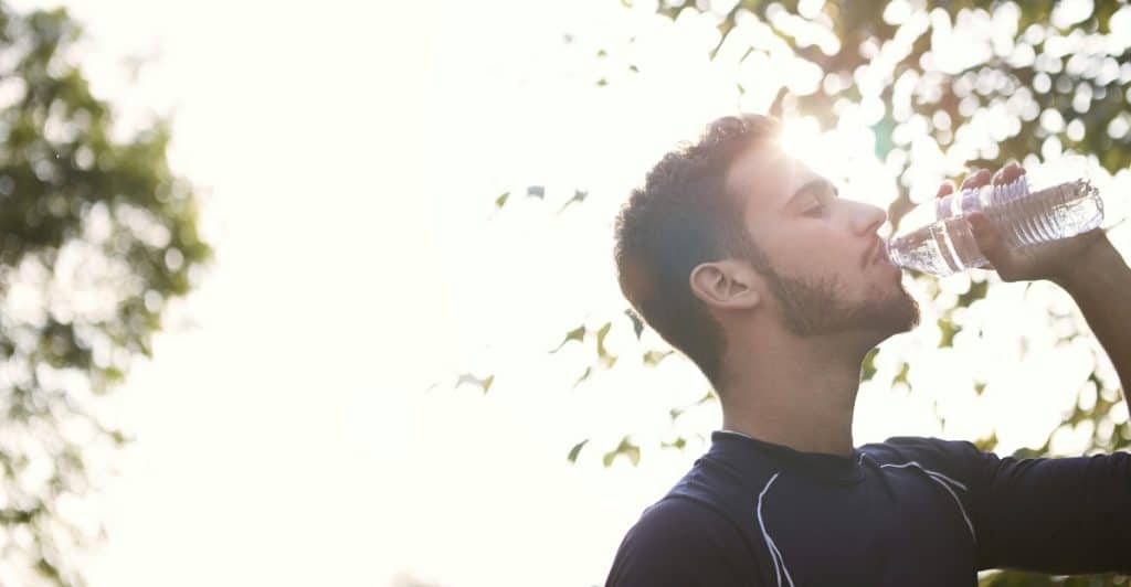 A bearded man in a black shirt drinking water from a plastic bottle with the sun shining behind him.
