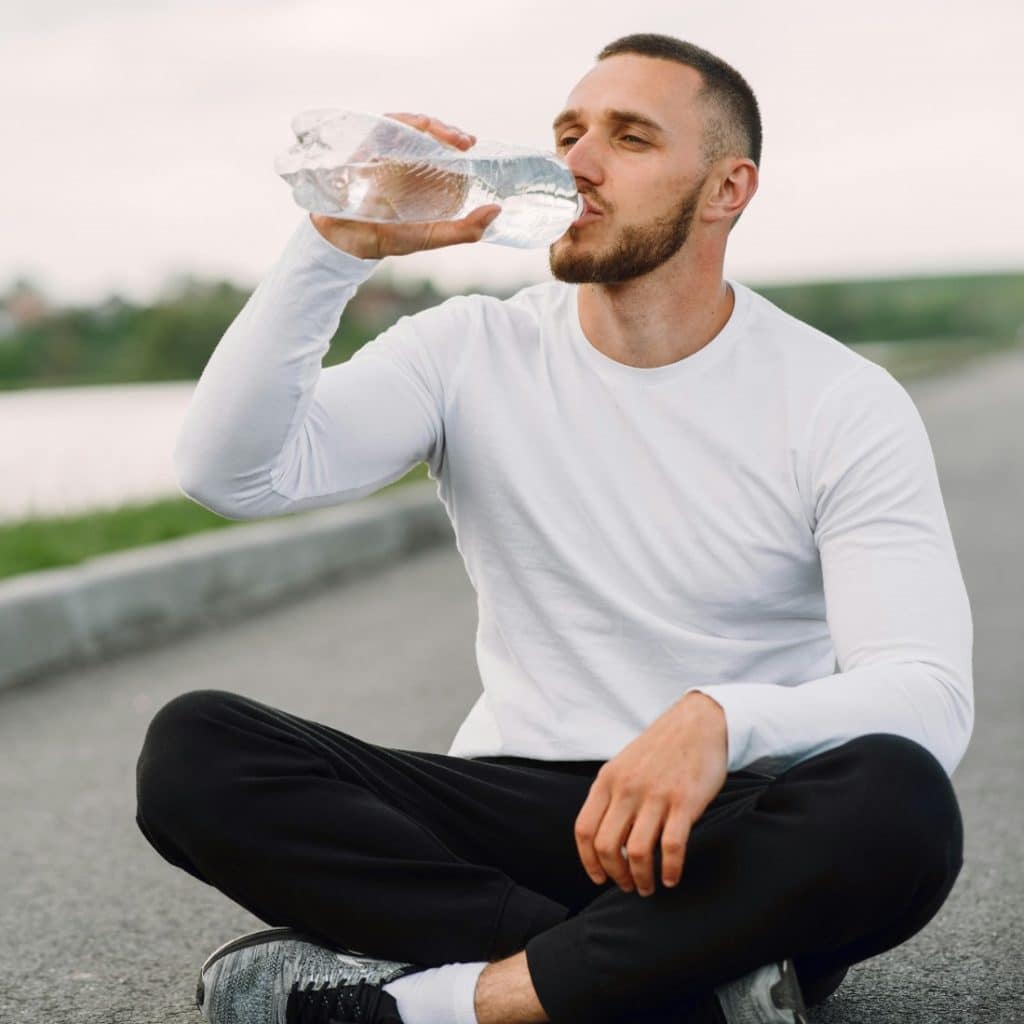 A man with a beard, wearing a white long-sleeve shirt and black pants, sits cross-legged on an asphalt road and drinks water from a plastic bottle.