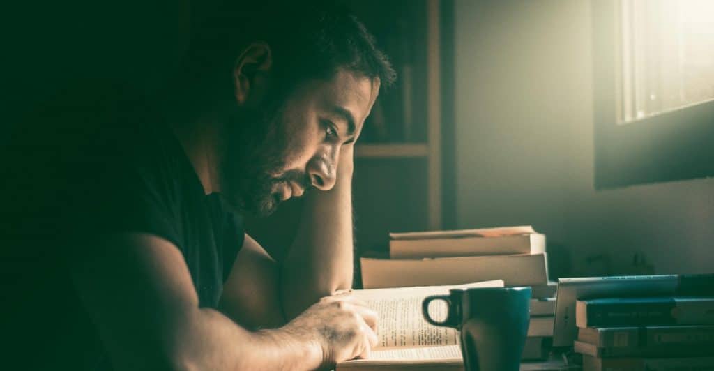 A man with a beard is reading a book at a table with a blue mug and stacks of books. He is looking down at the book with a focused expression, and the lighting is dim.