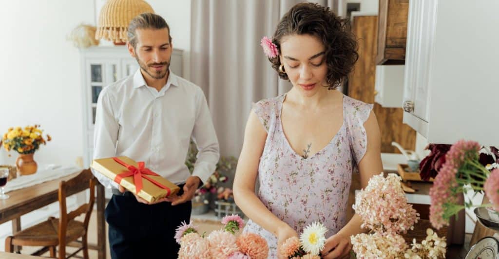 A man holding a wrapped gift while a woman arranges flowers at a table.