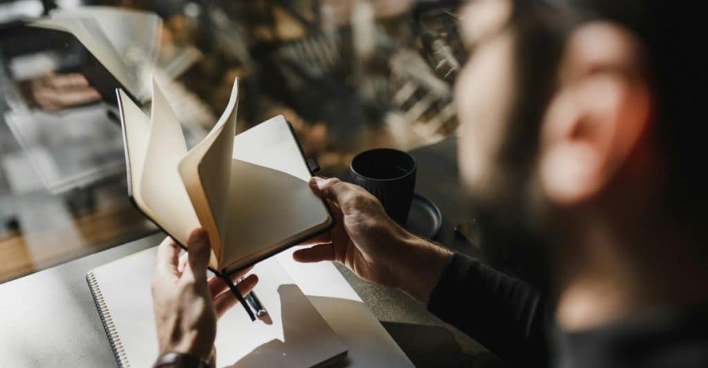 A man is writing in a notebook at a table in a cafe, with a coffee cup and another notebook visible.