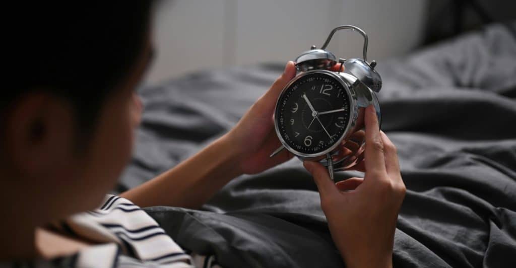 A person in bed holding an analog alarm clock, with the clock face clearly visible, suggesting the start of the day.