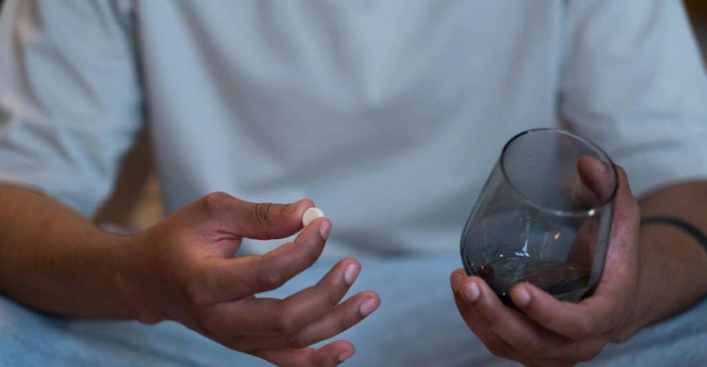 A close-up of a person's hands holding a white pill and a glass of water.