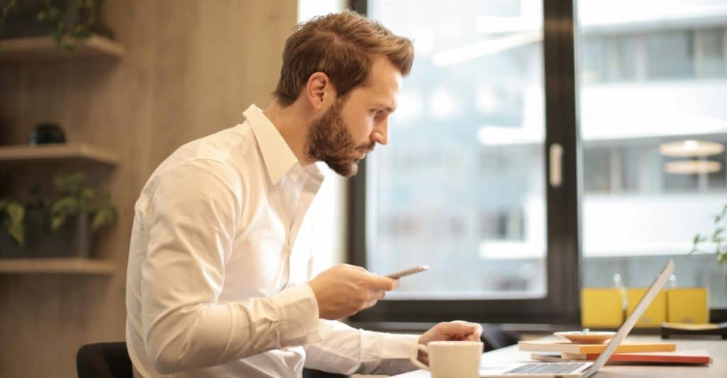 A man with a beard, wearing a white shirt, sits at a desk looking at his smartphone while working on a laptop.