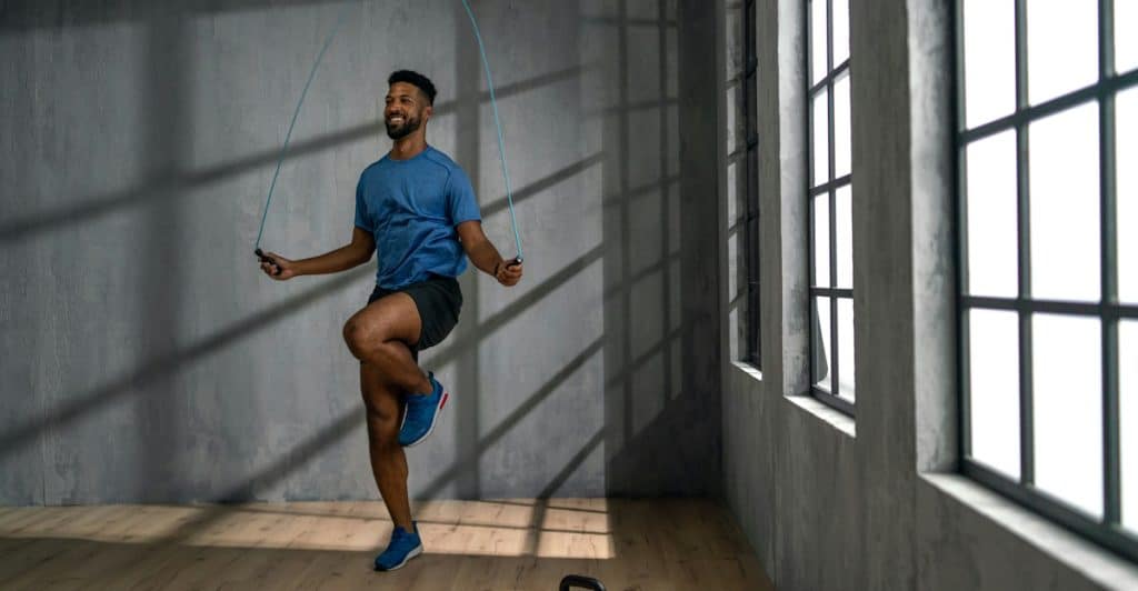 A man with dark skin, wearing a blue t-shirt and shorts, jumping rope in a room with gray walls, large windows, and a kettlebell on the floor.