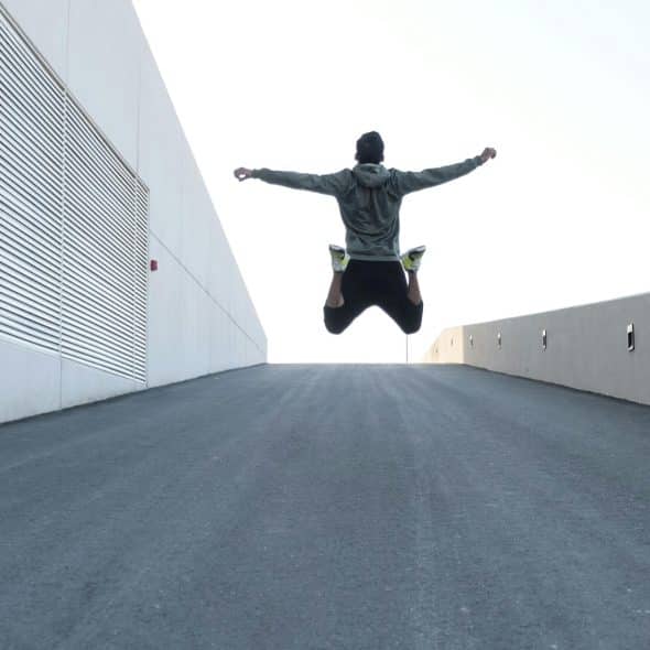 A man wearing athletic clothing is captured mid-jump with arms outstretched against a bright sky and modern architecture.