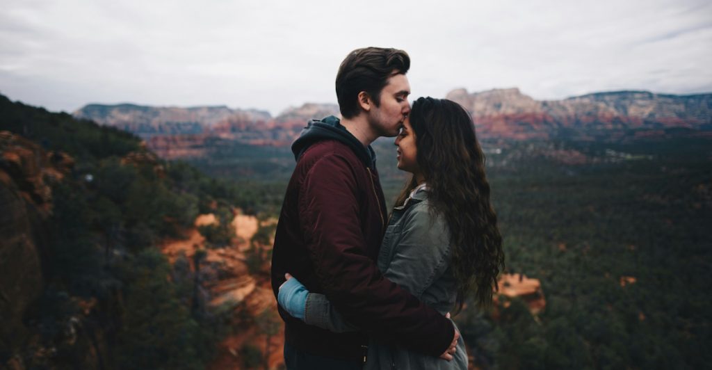 A man kissing a woman's forehead on a rocky overlook, with a mountainous landscape in the background.